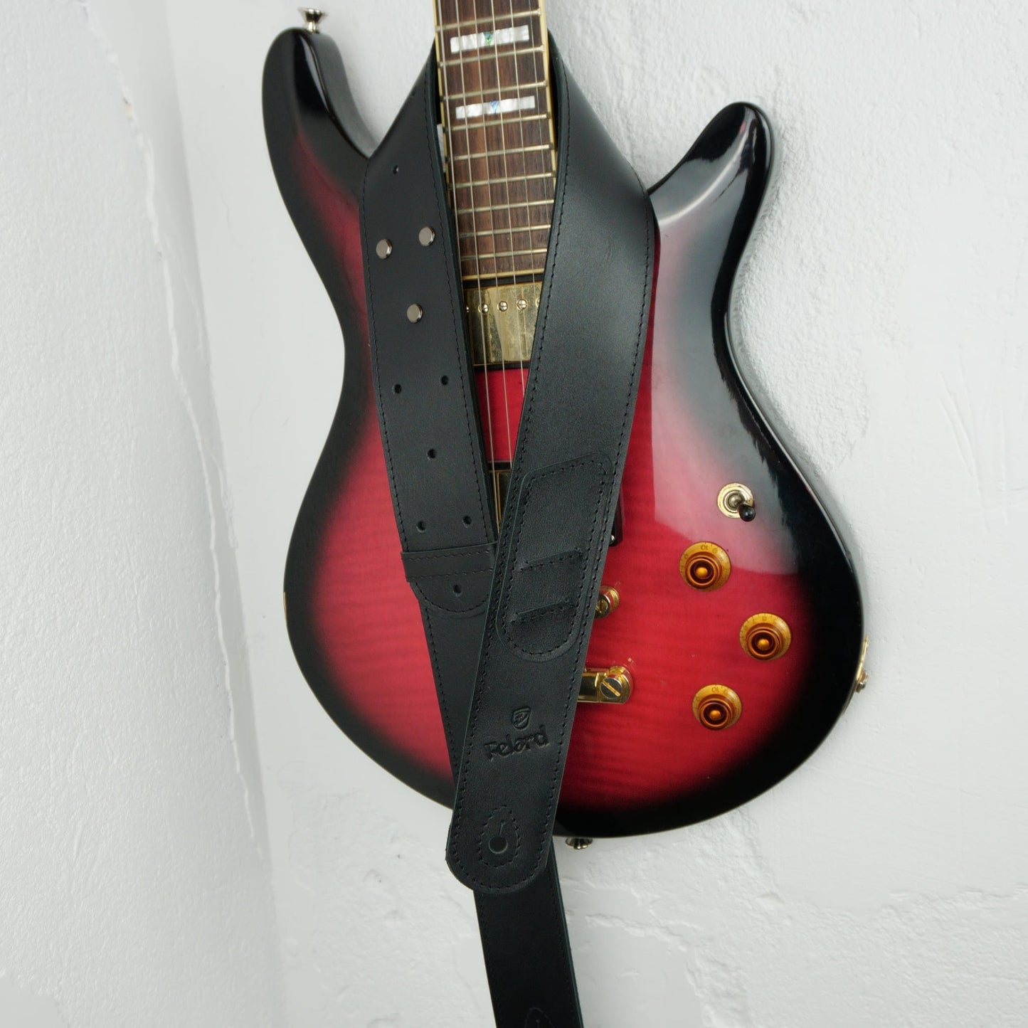 A black leather electric guitar strap with a visible 'Felard' logo, hanging over the body of a red-to-black gradient electric guitar against a white wall.