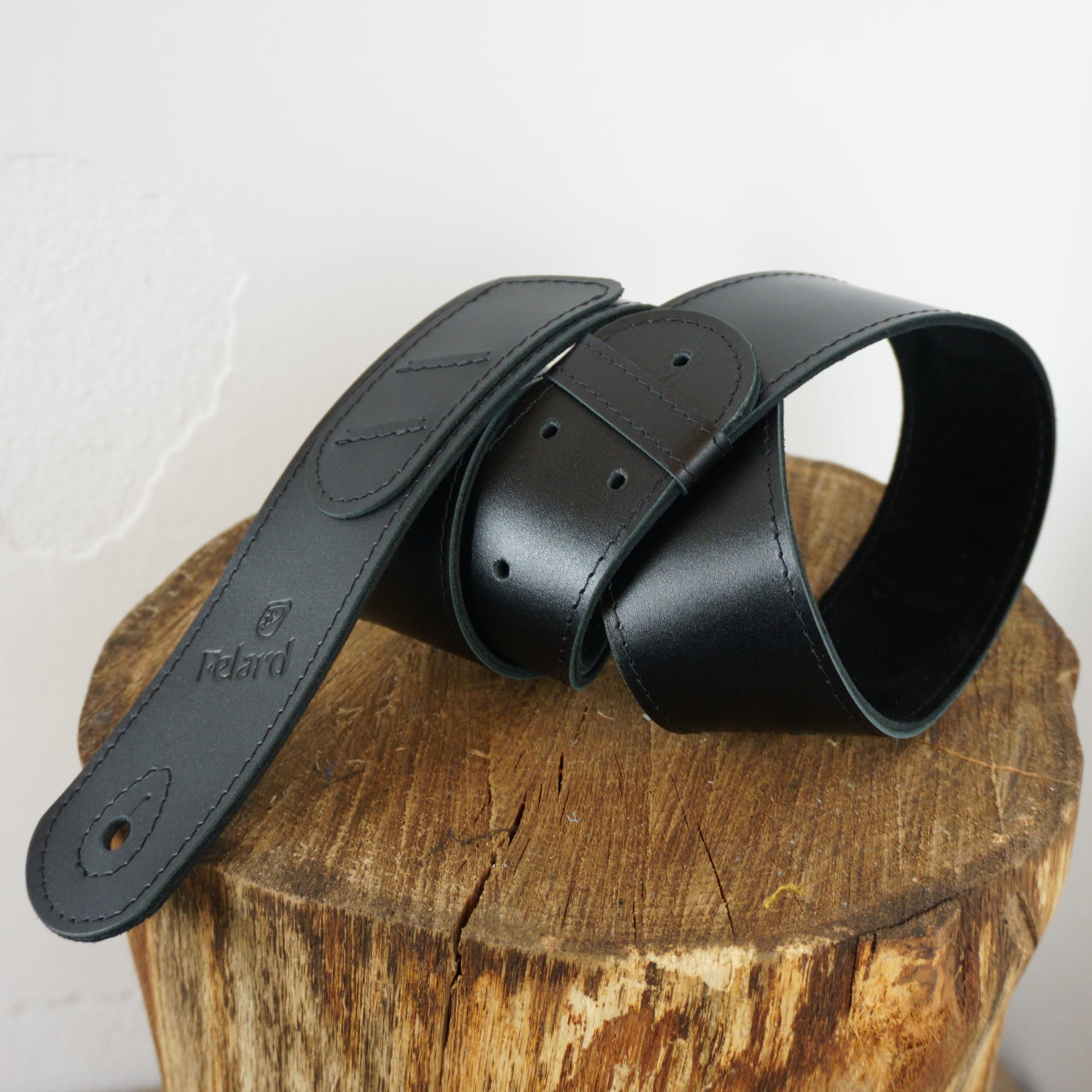 A black leather guitar strap on a rustic wooden log against a plain white wall.