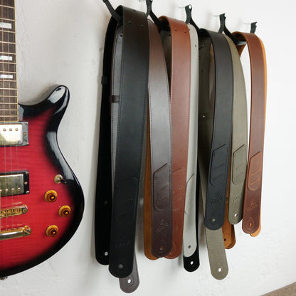 Leather guitar straps in various colors (black, brown, silver, dark gray, olive green, tan) hanging from black hooks on a white wall next to a red electric guitar.