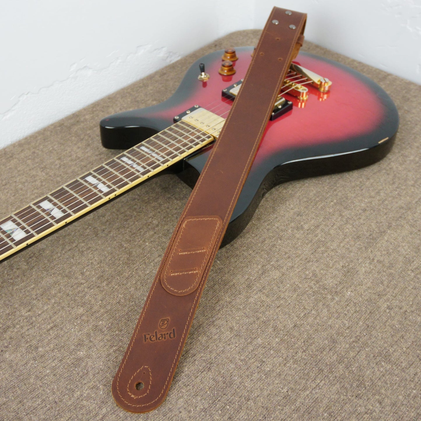 A brown leather guitar strap with an embossed 'Felard' logo, laid diagonally across a red and black electric guitar on a textured gray carpet.
