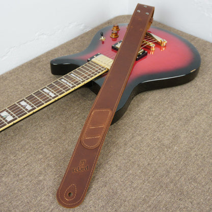 A brown leather guitar strap with an embossed 'Felard' logo, laid diagonally across a red and black electric guitar on a textured gray carpet.