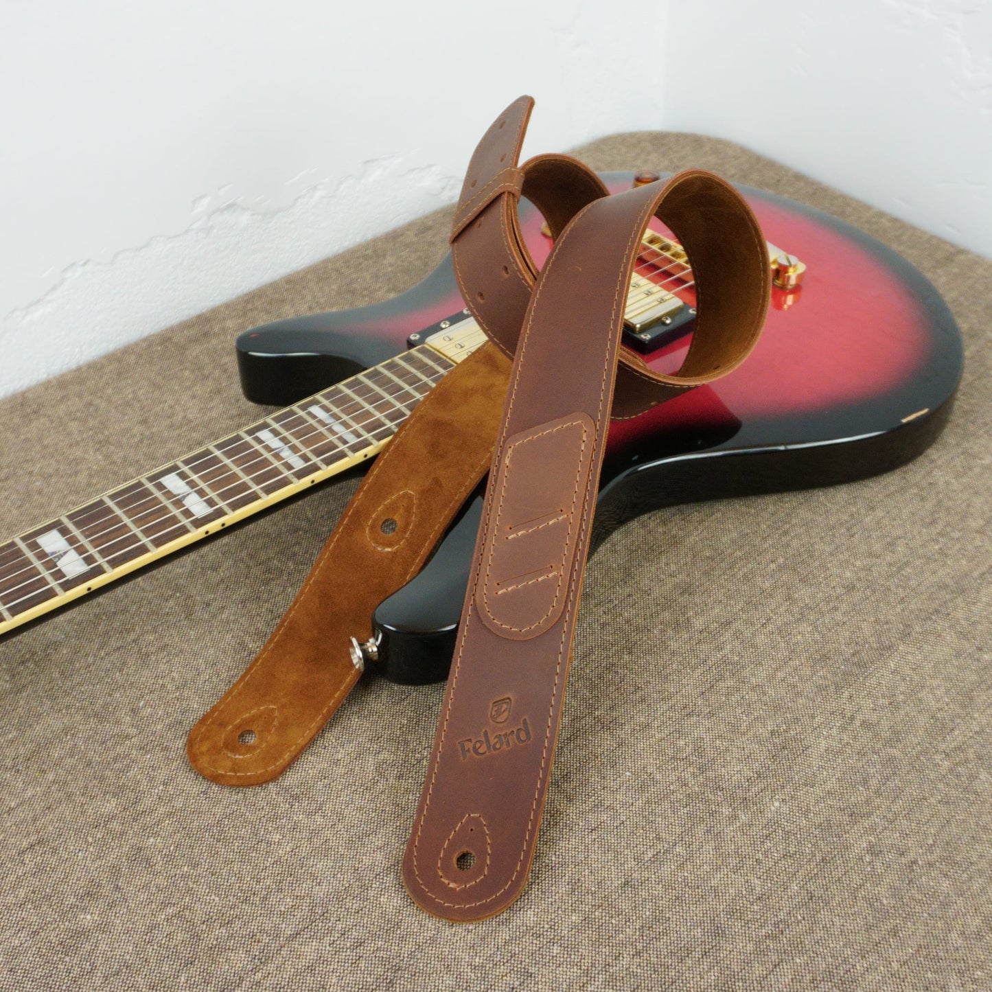 A brown leather guitar strap with a suede-like section and metal hardware, draped over the body of a red and black electric guitar on a textured carpet.