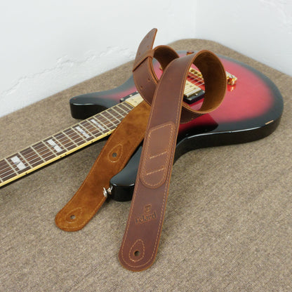 A brown leather guitar strap with a suede-like section and metal hardware, draped over the body of a red and black electric guitar on a textured carpet.