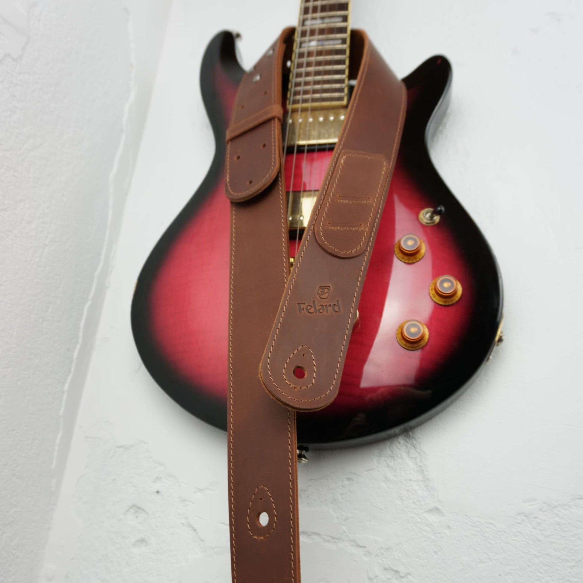 A brown leather guitar strap with a visible 'Felard' logo, draped over a red-to-black gradient electric guitar against a white textured wall.