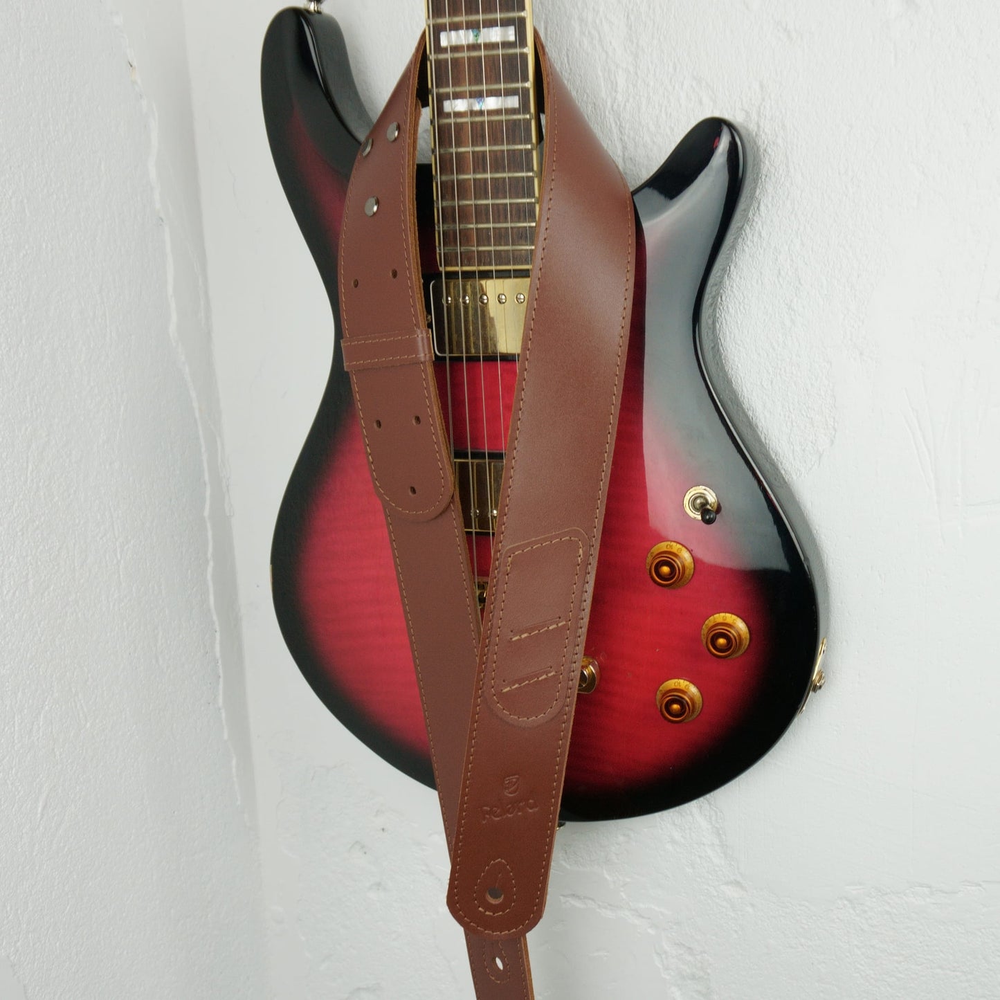 A brown leather guitar strap hanging vertically in front of a red-to-black sunburst electric guitar against a white wall.