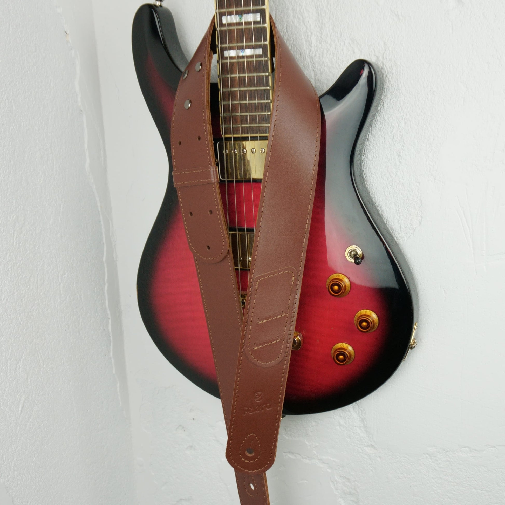 A brown leather guitar strap hanging vertically in front of a red-to-black sunburst electric guitar against a white wall.