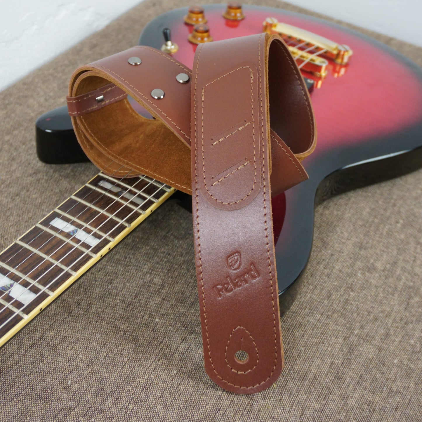A brown leather guitar strap with metal rivets and an embossed 'Felard' logo, draped over a red and black electric guitar on a textured beige surface.