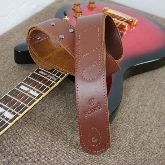 A brown leather guitar strap with metal rivets and an embossed 'Felard' logo, draped over a red and black electric guitar on a textured beige surface.