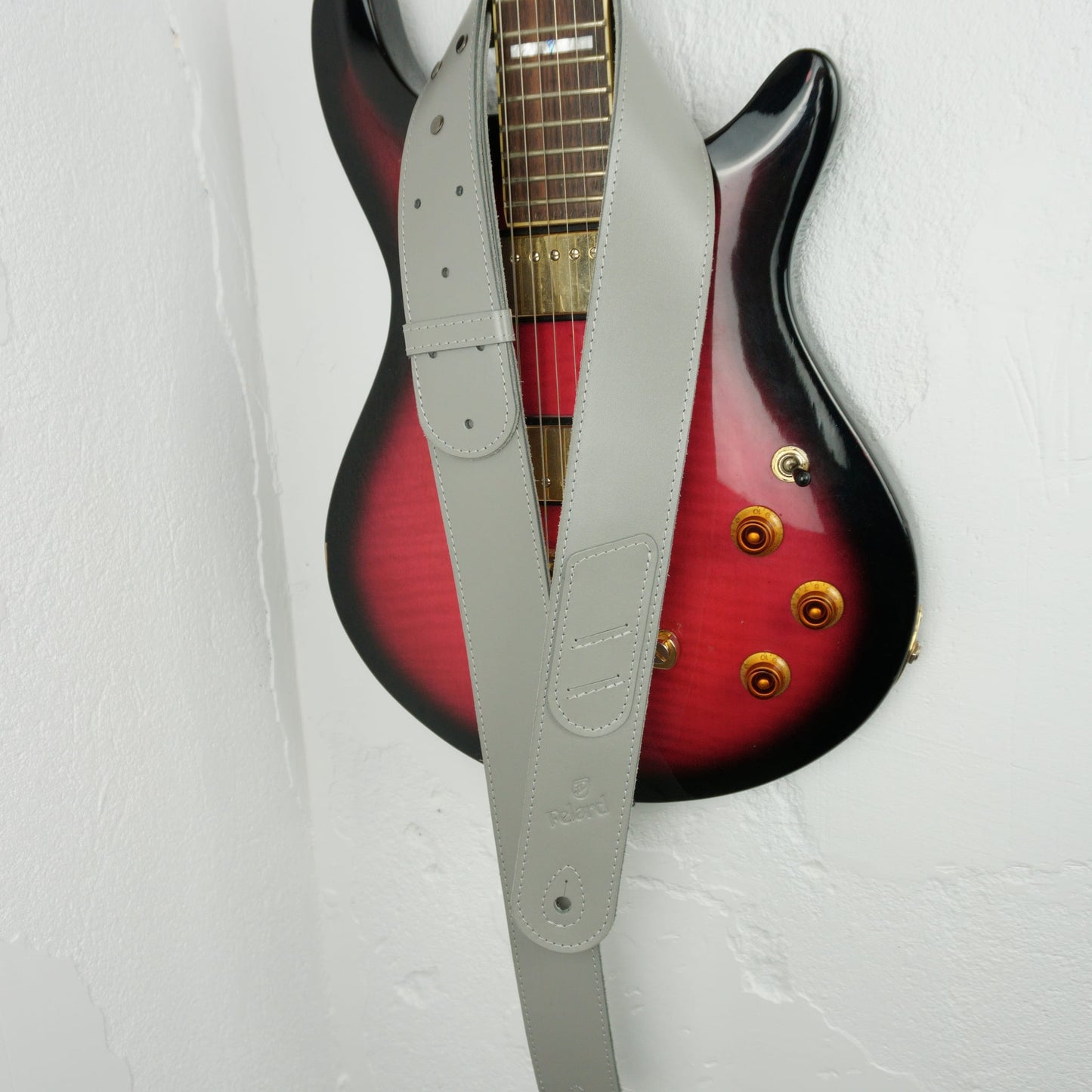 A light gray guitar strap hanging vertically in front of a red-to-black gradient electric guitar against a white textured wall.