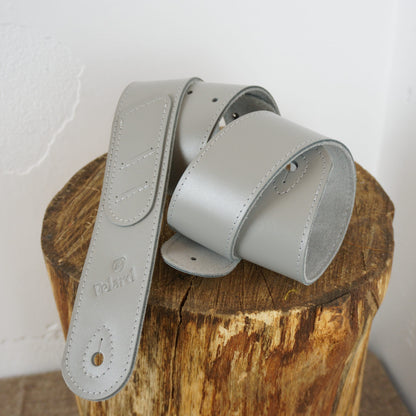A light gray leather guitar strap resting on a rustic wooden log stump against a white wall. The brand name 'Felard' is embossed on the strap.