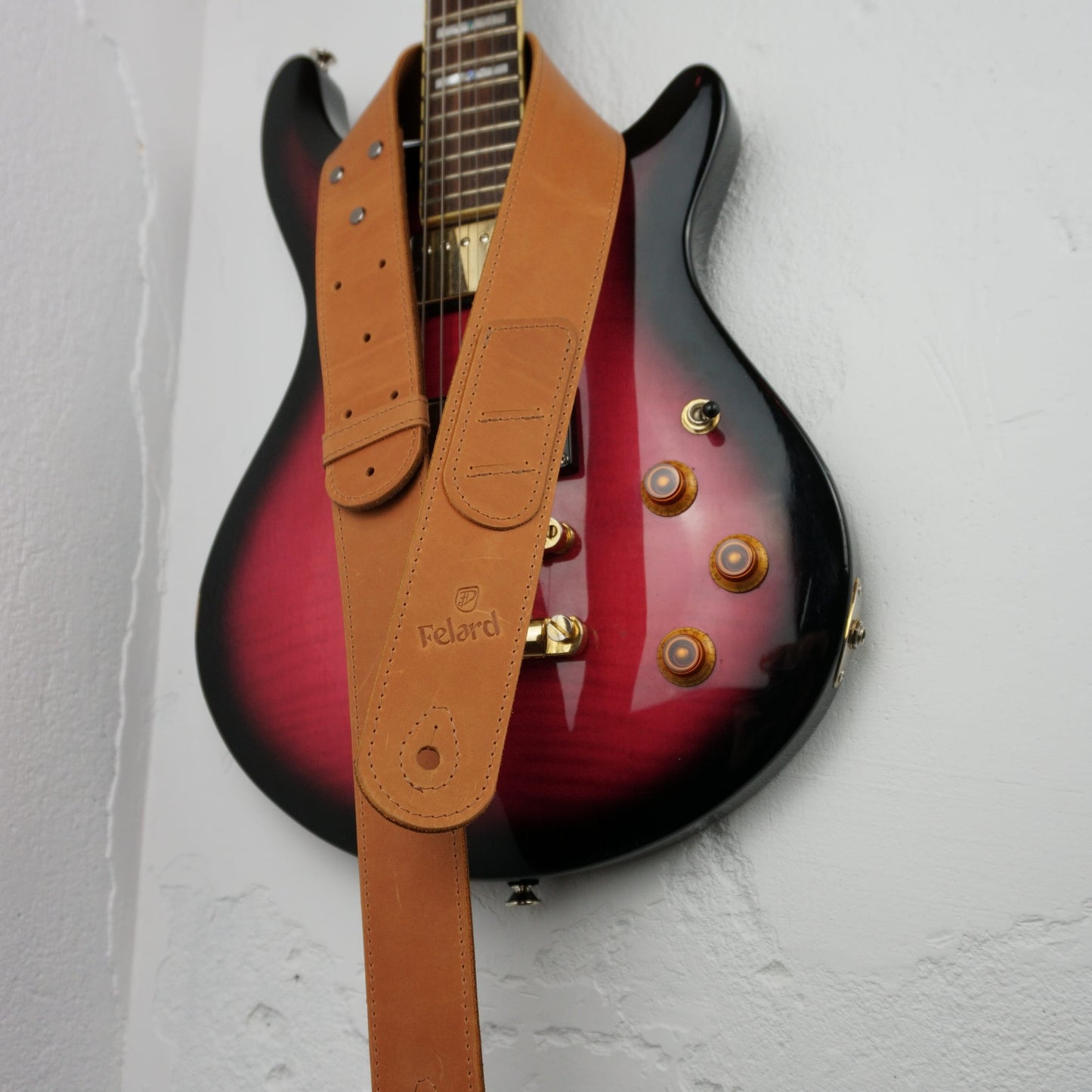 A tan leather guitar strap with the brand name 'Felard' embossed on it, draped over a red-to-black gradient electric guitar against a white textured wall.