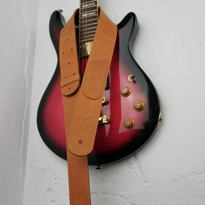 A tan leather guitar strap with the brand name 'Felard' embossed on it, draped over a red-to-black gradient electric guitar against a white textured wall.