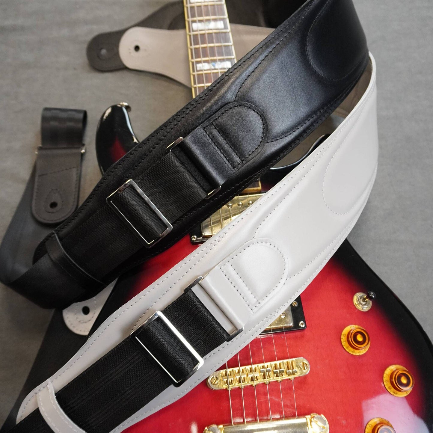 A close-up of a guitar straps laid across a red electric guitar on a gray surface. The strap is shown in two colorways: a black leather version with silver buckles and a gray leather version with silver buckles.