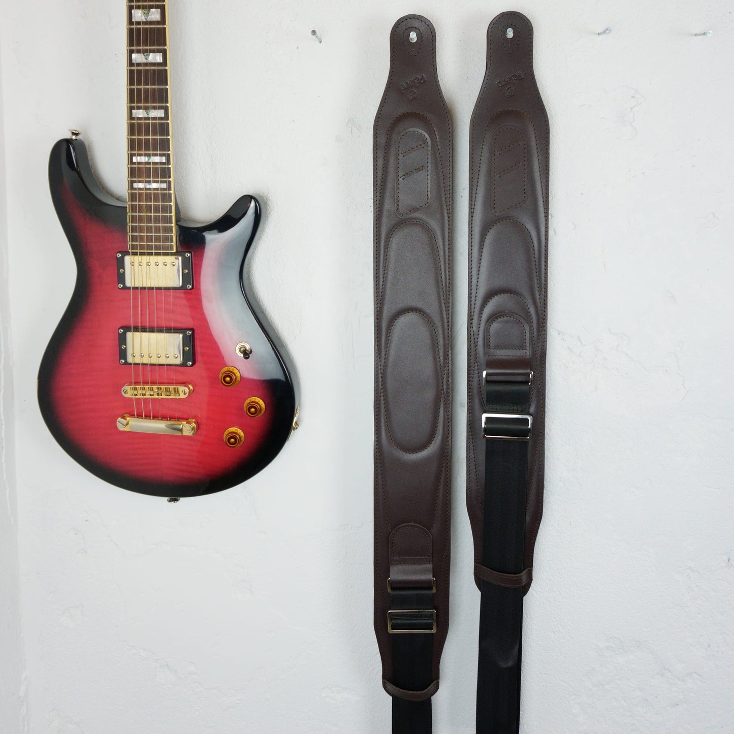 Two dark brown guitar straps displayed vertically next to a red and black electric guitar on a white wall.
