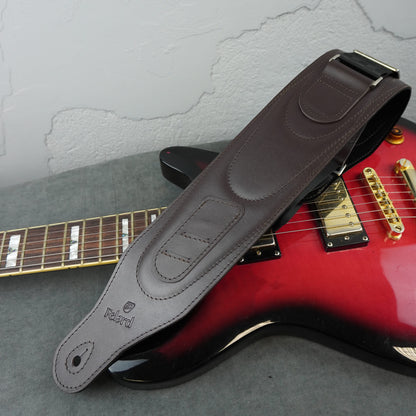 A dark brown leather guitar strap with 'Felard' branding, draped over a red electric guitar on a gray textured surface against a light gray wall.