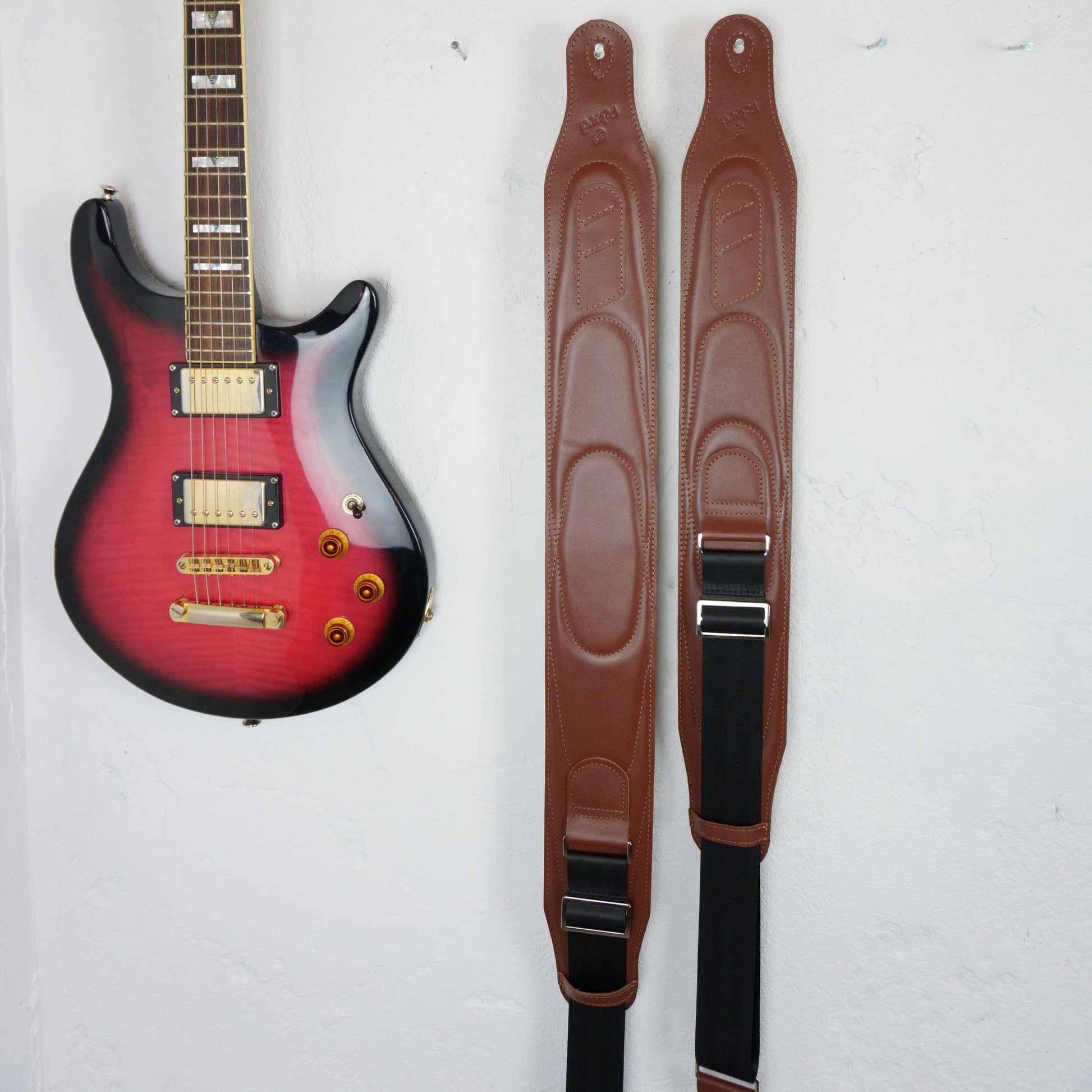 Two brown guitar straps hanging vertically on a white wall next to a red and black electric guitar.