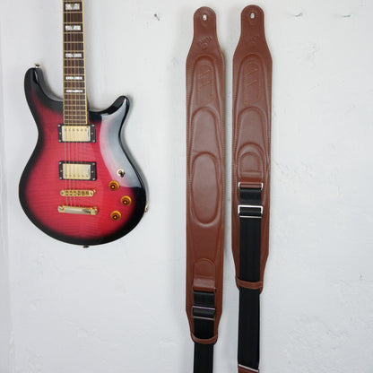 Two brown guitar straps hanging vertically on a white wall next to a red and black electric guitar.