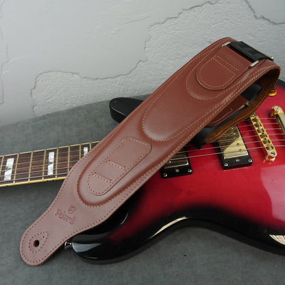 A brown smooth leather guitar strap with 'Felard' branding, laid diagonally across a red electric guitar on a gray surface.