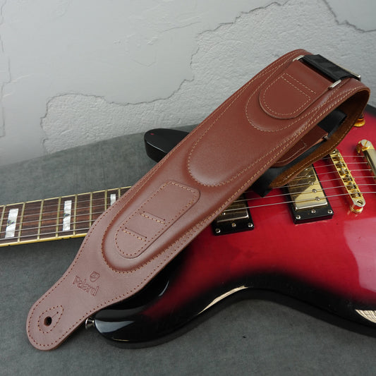 A brown smooth leather guitar strap with 'Felard' branding, laid diagonally across a red electric guitar on a gray surface.