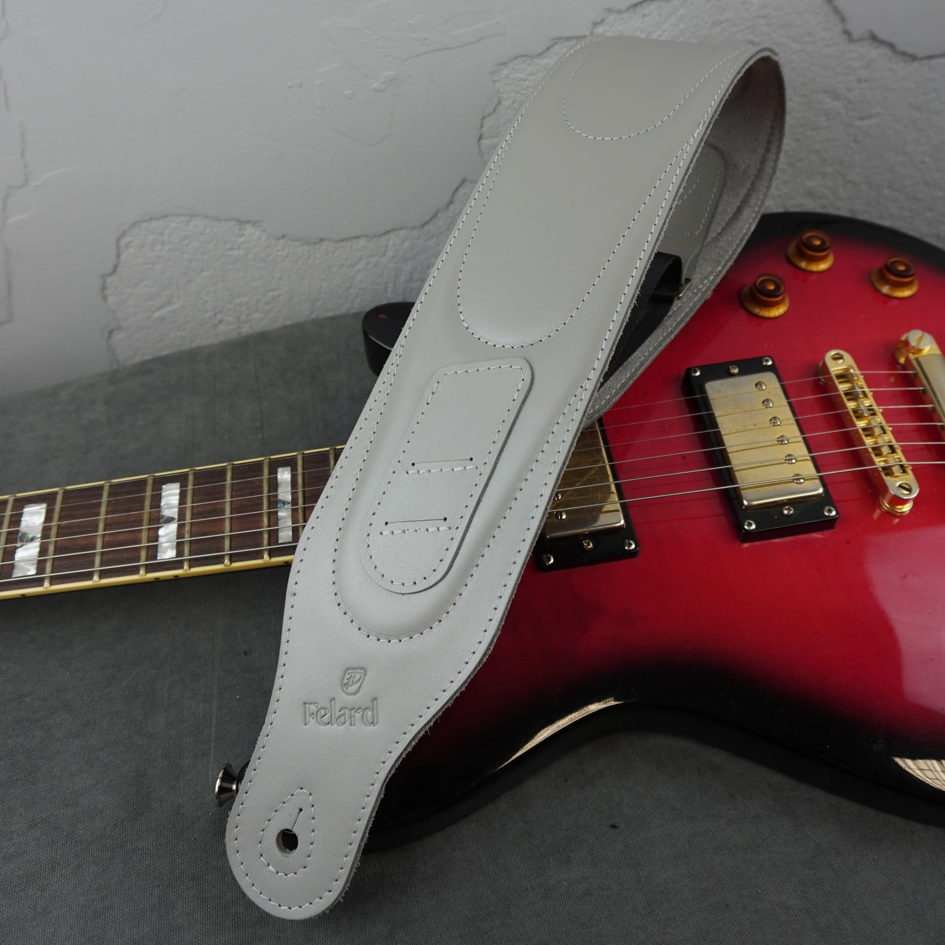 A light gray leather guitar strap with the Felard logo, draped over a red electric guitar on a gray surface against a textured gray wall.