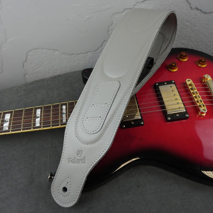 A light gray leather guitar strap with the Felard logo, draped over a red electric guitar on a gray surface against a textured gray wall.
