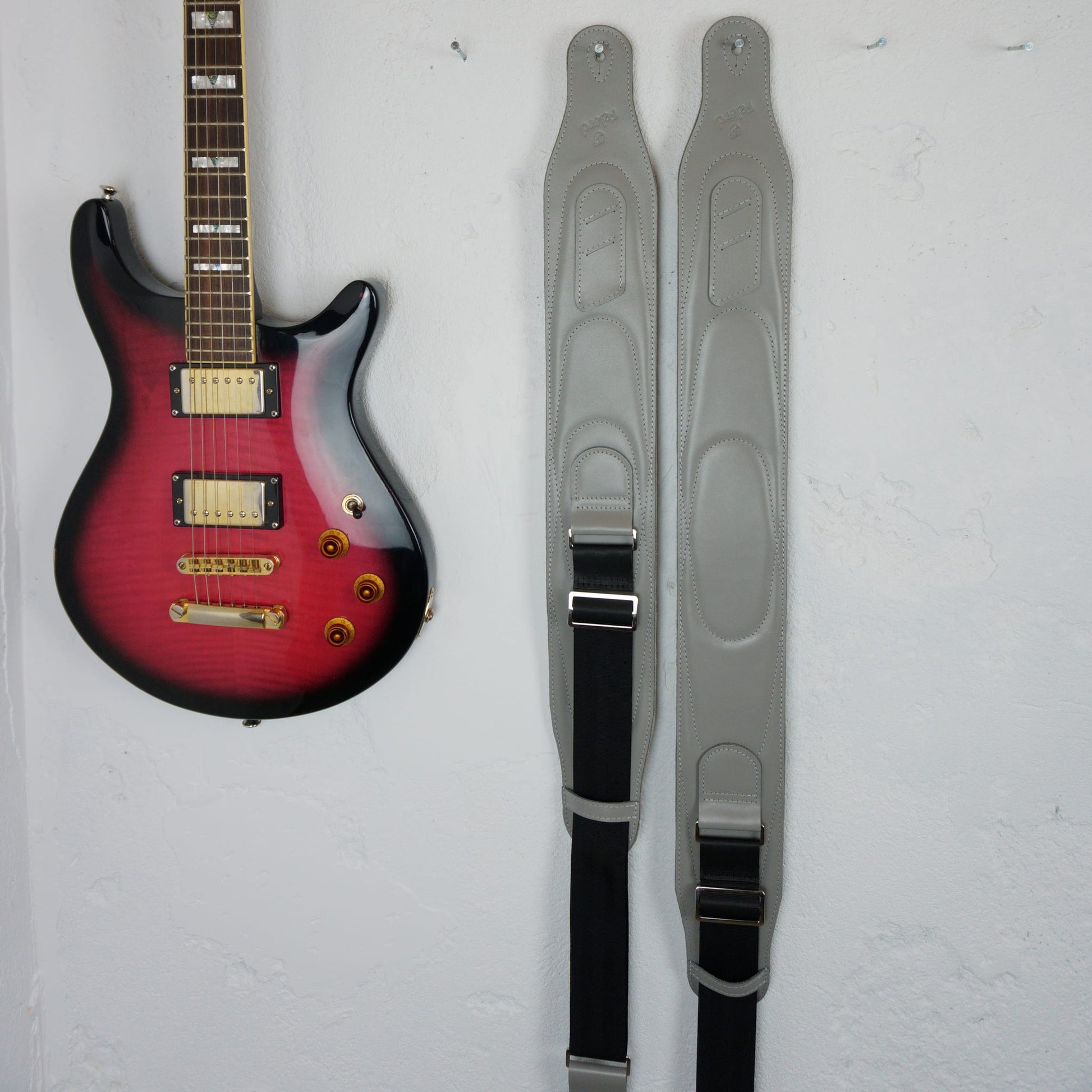 A pair of gray guitar straps hanging vertically on a white wall next to a red and black electric guitar.