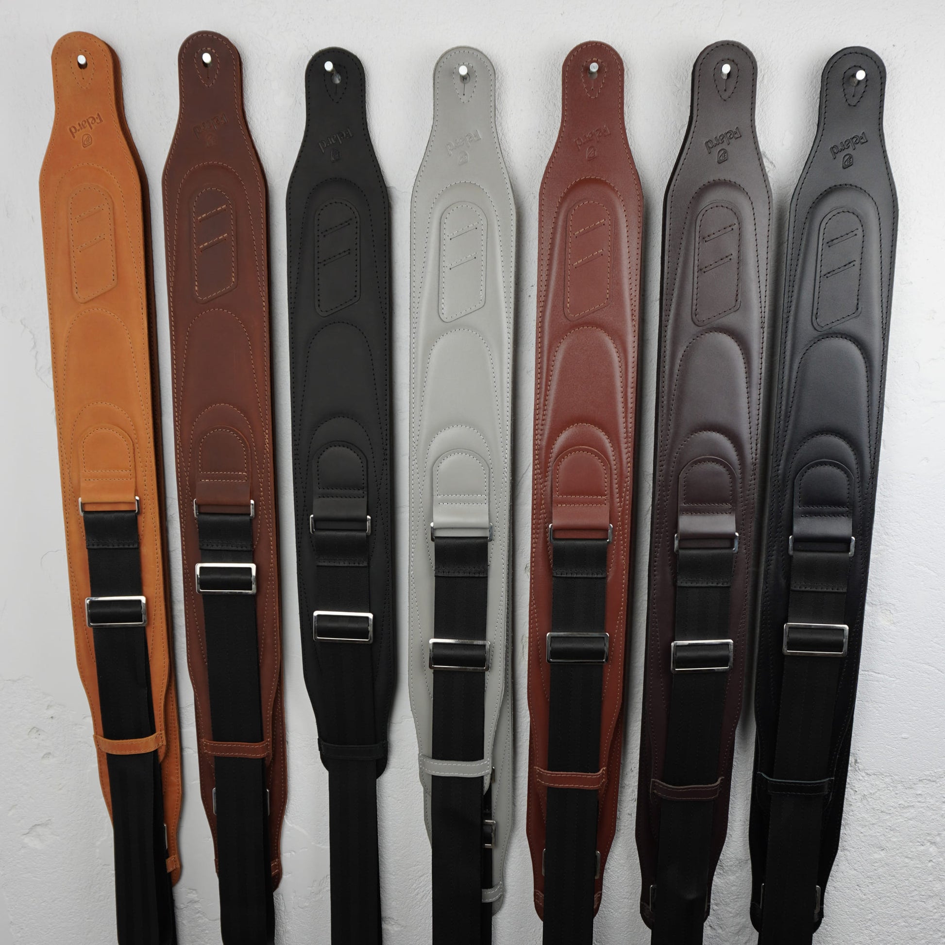 Guitar straps in different colors (tan, brown, black, light gray, reddish brown, dark brown) are lined up vertically against a white textured wall.