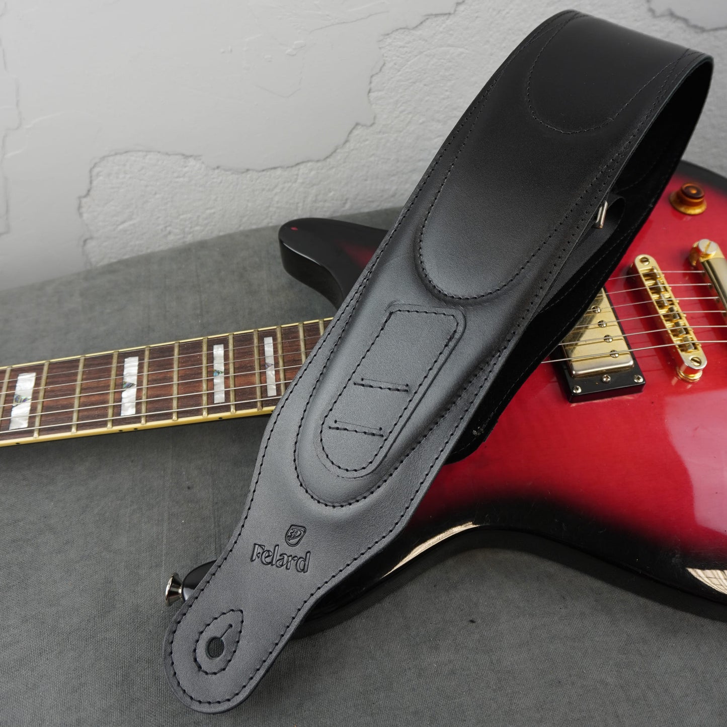 A black leather guitar strap with 'Felardi' branding over a red electric guitar on a gray surface against a textured white wall.