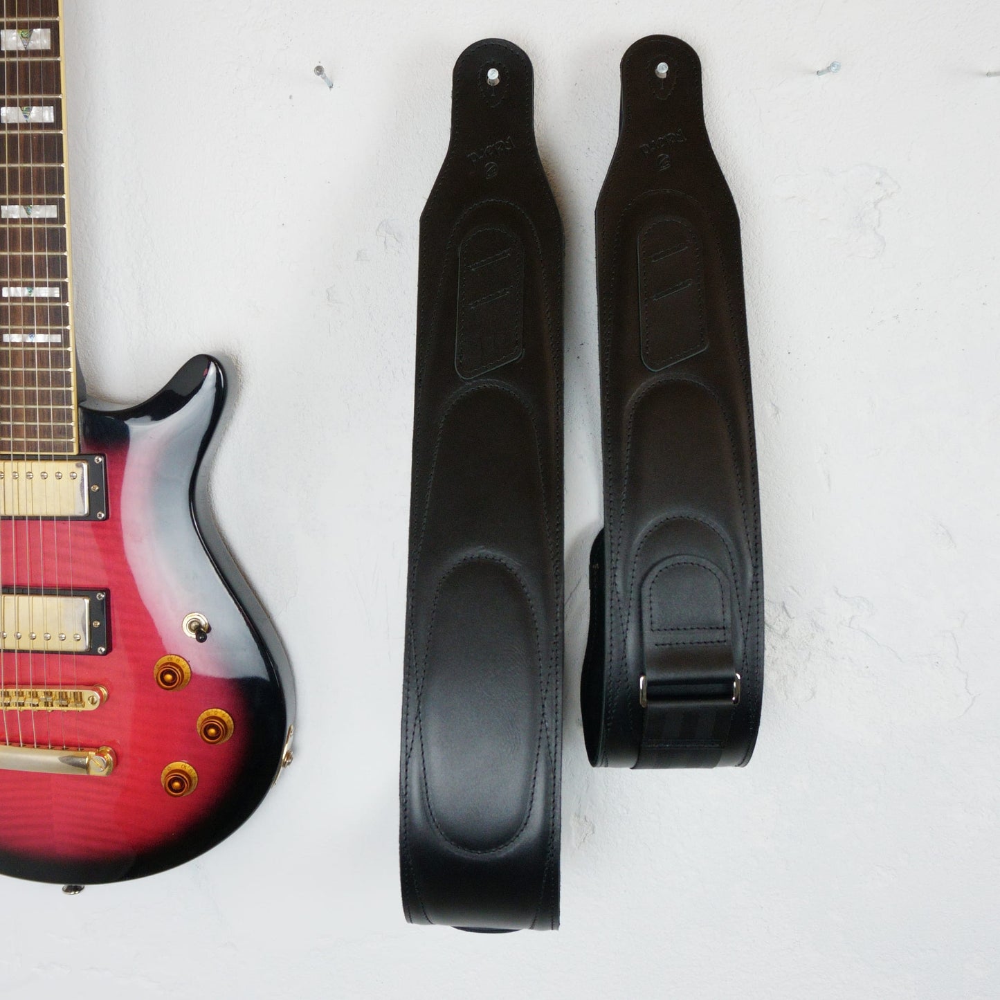 Two black guitar straps displayed flat on a white background, positioned next to the lower portion of a red-to-black gradient electric guitar.