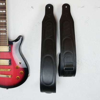 Two black guitar straps displayed flat on a white background, positioned next to the lower portion of a red-to-black gradient electric guitar.