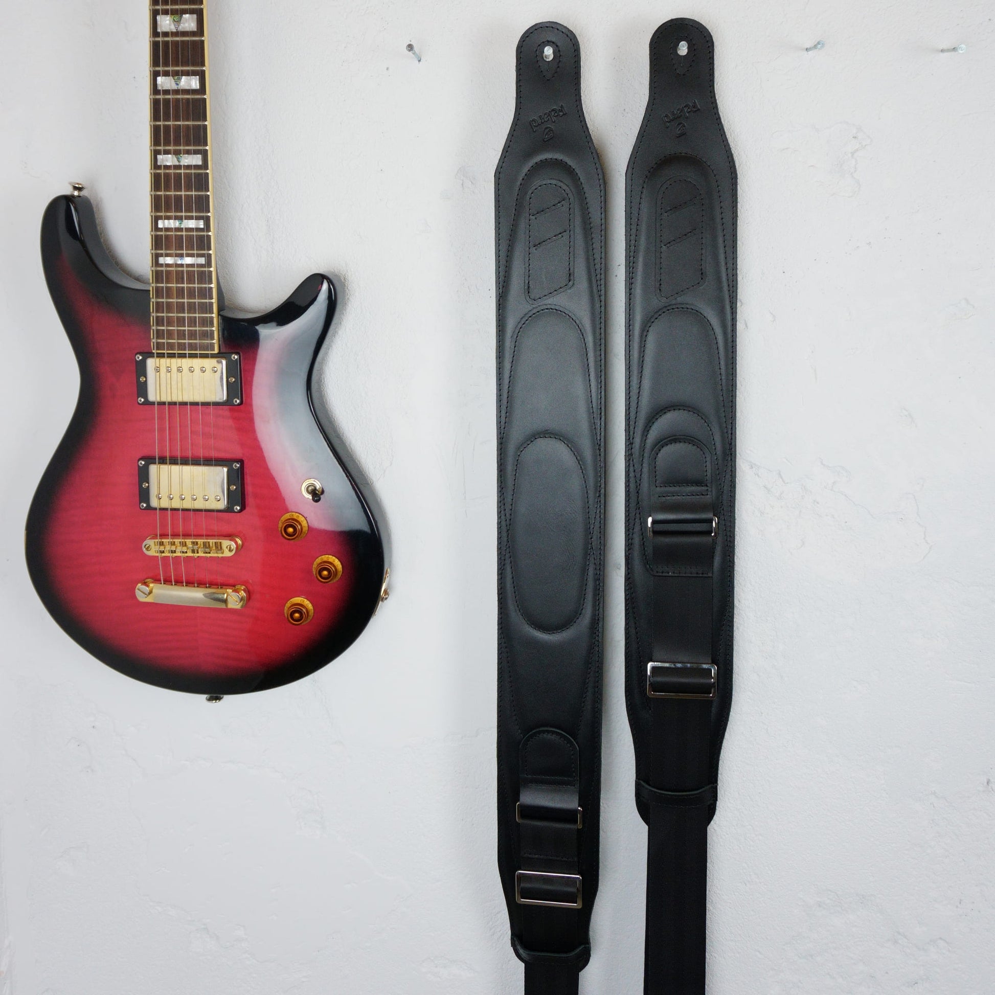 Two black guitar straps displayed vertically next to a red-to-black gradient electric guitar on a white textured wall.