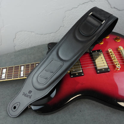 A black leather electric guitar strap with 'Felard' branding over a red electric guitar on a gray surface against a textured white wall.