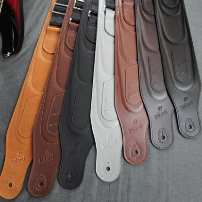 Leather guitar straps in different colors arranged diagonally on a gray fabric background. The straps are tan, reddish-brown, black, off-white, light brown, and dark gray.