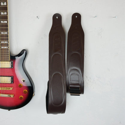 A dark brown guitar straps with stitched oval patterns and a metal buckle, displayed next to a red-to-black gradient electric guitar on a textured white background.