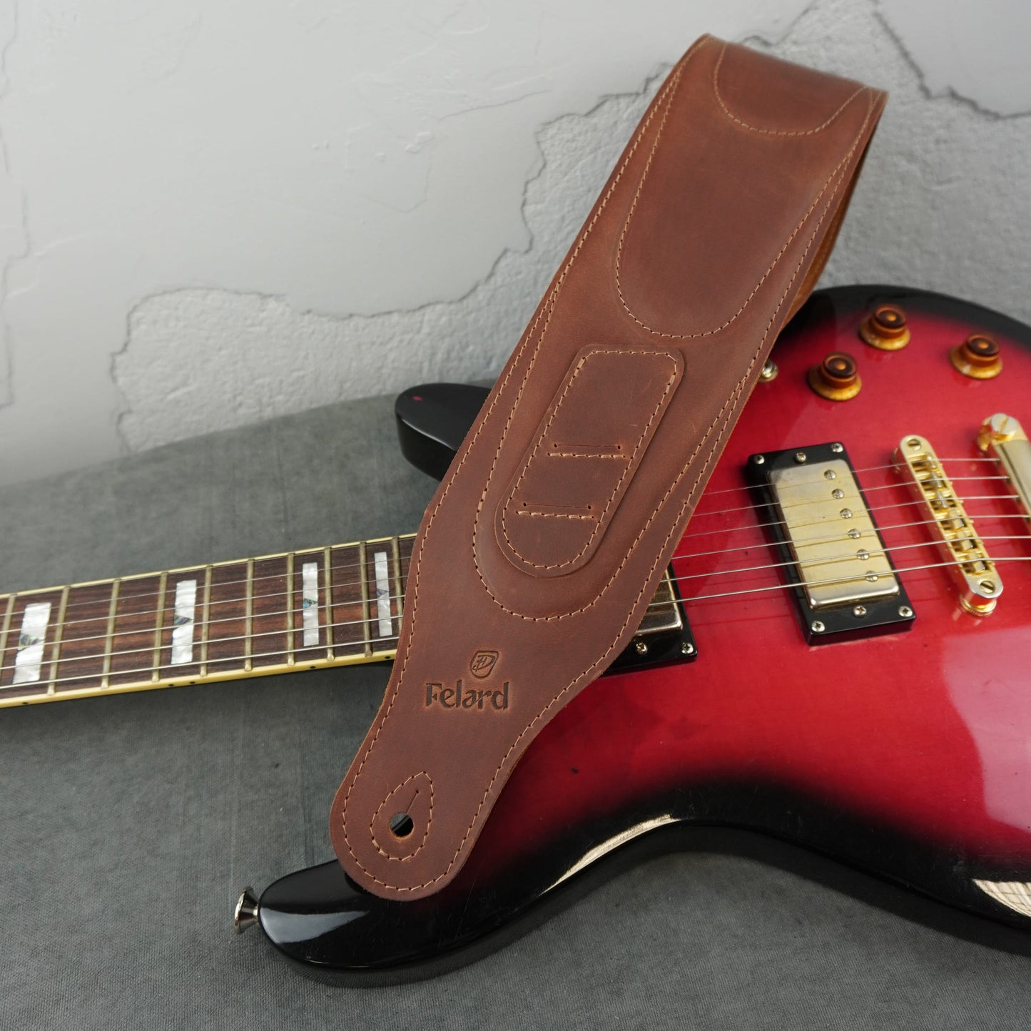 A brown leather guitar strap with the brand name 'Felard' embossed on it, draped over a red electric guitar against a gray textured background.