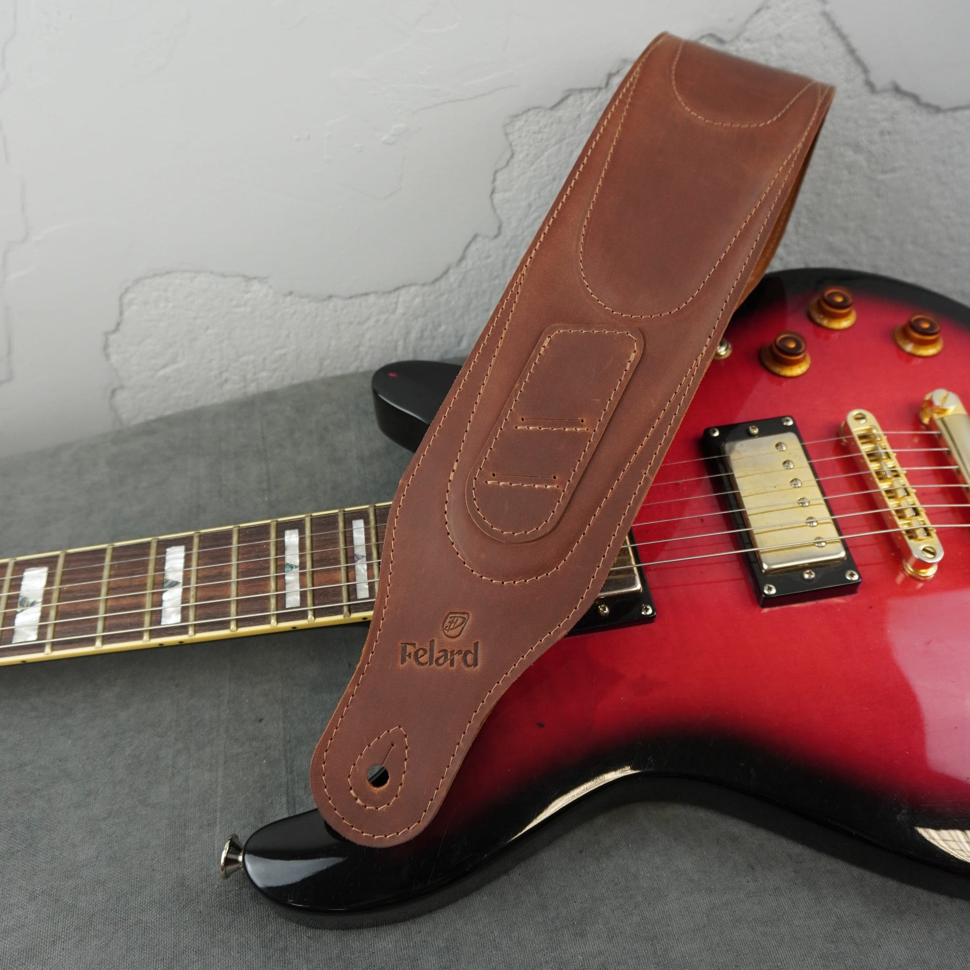 A brown leather guitar strap with the brand name 'Felard' embossed on it, draped over a red electric guitar against a gray textured background.