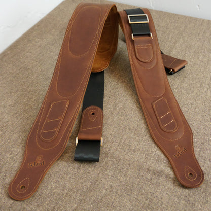 A pair of brown leather Felard guitar straps laid flat on a textured beige fabric surface, with black webbing and brass-tone buckles visible.