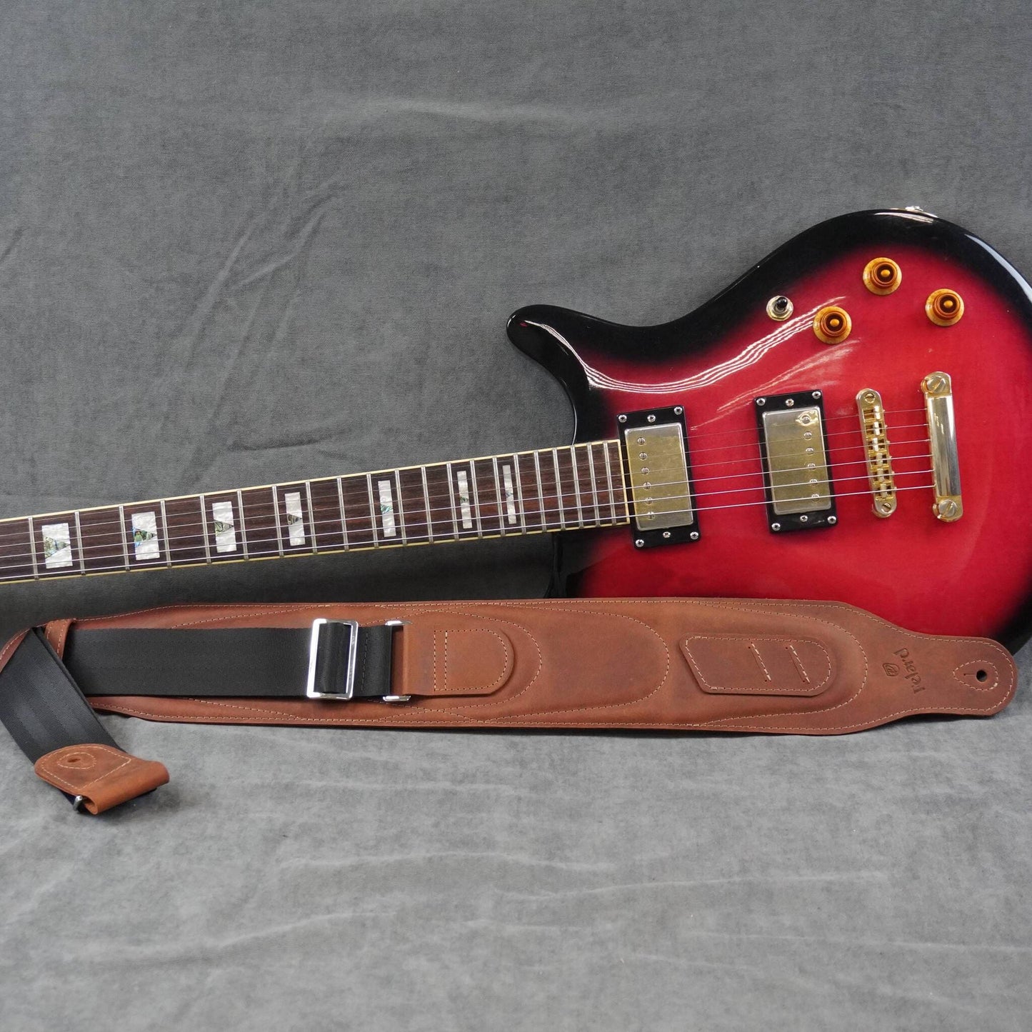A red electric guitar with a brown leather guitar strap laid out on a gray fabric background.