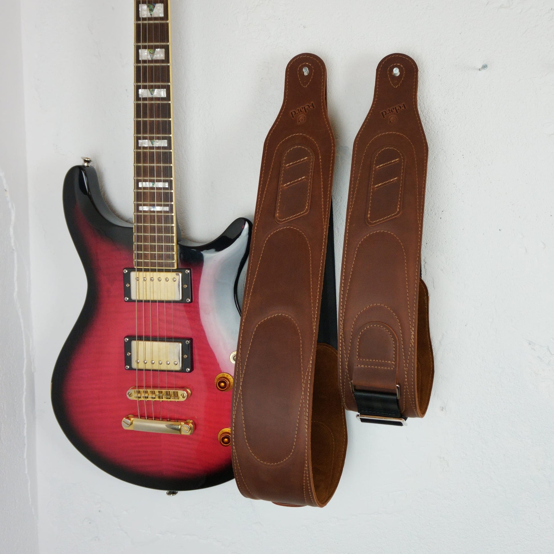 A brown leather electric guitar straps displayed next to a red-to-black gradient guitar against a plain white wall.