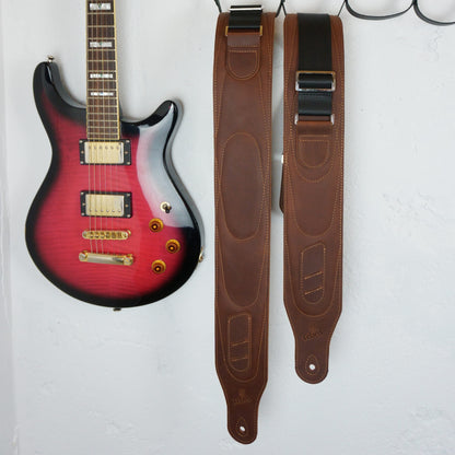 A pair of brown leather guitar straps hanging on a white wall next to a red-to-black gradient electric guitar.
