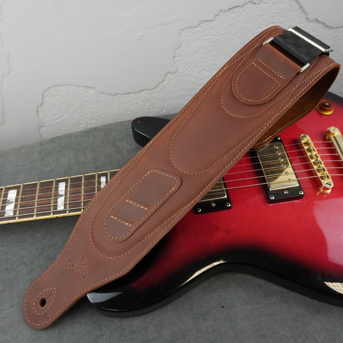 A brown leather guitar strap with silver metal hardware, laid diagonally across a red and black electric guitar on a gray surface.