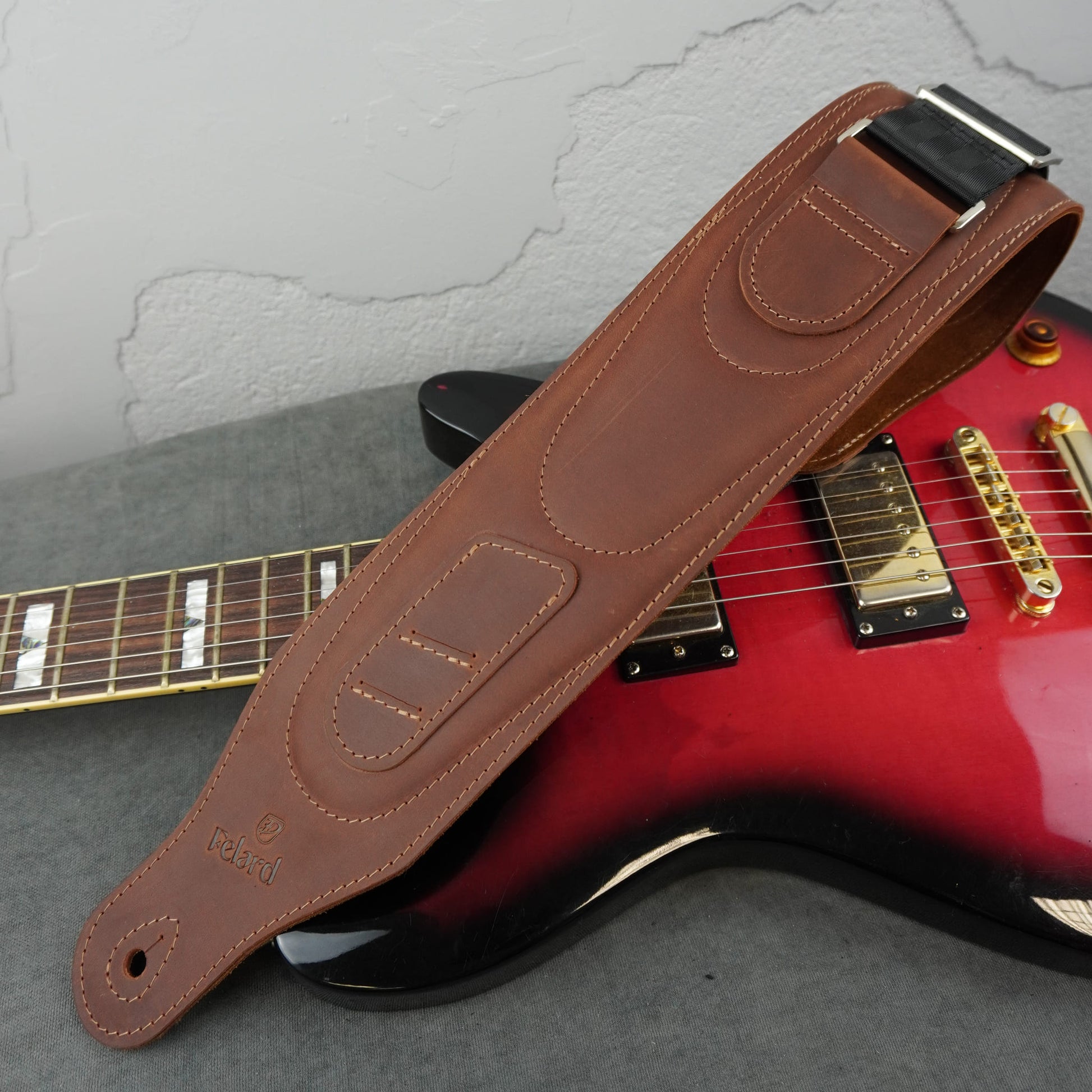 A brown leather guitar strap with silver metal hardware, laid diagonally across a red and black electric guitar on a gray surface.