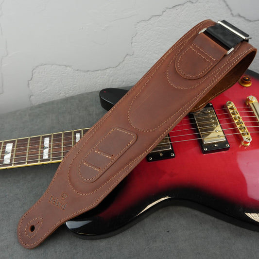 A brown leather guitar strap with silver metal hardware, laid diagonally across a red and black electric guitar on a gray surface.