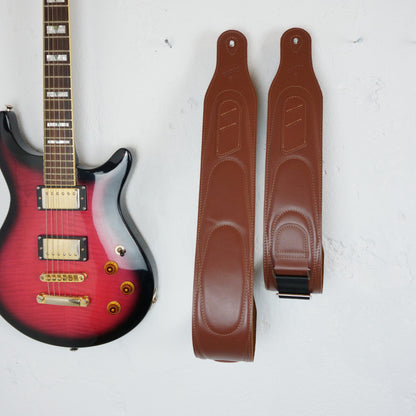 A pair of brown leather guitar straps displayed vertically next to a red-to-black gradient electric guitar on a white textured background.
