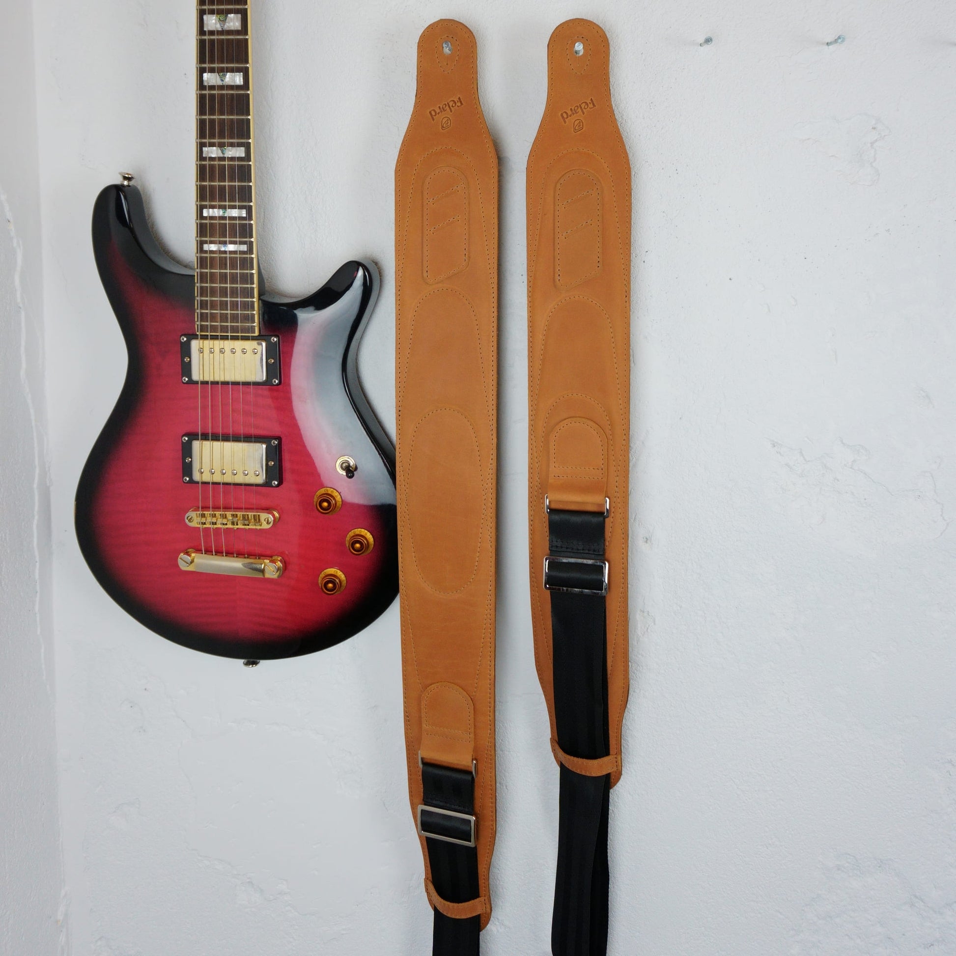 Two tan leather guitar straps with black webbing and silver metal buckles, displayed vertically next to a red-to-black gradient electric guitar against a white textured wall.