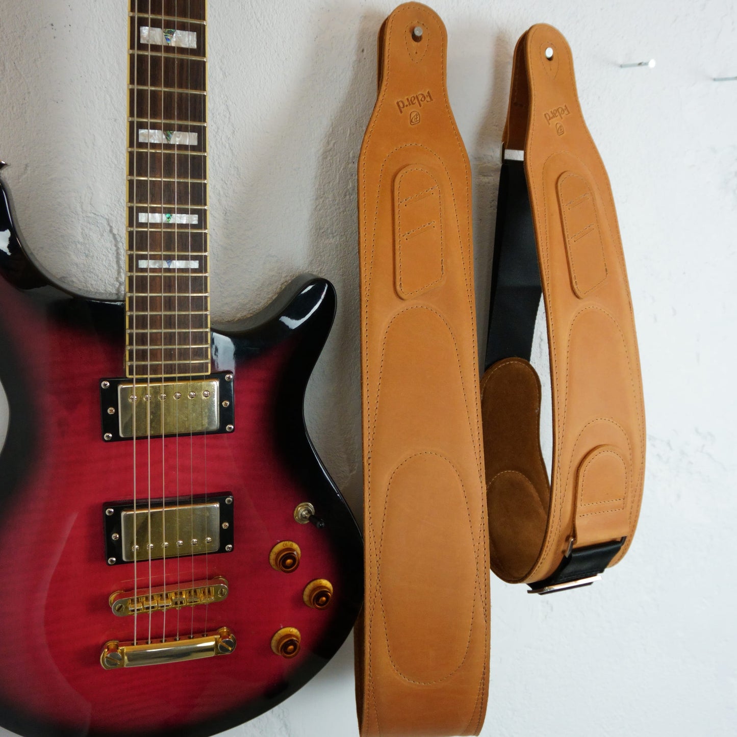 A tan leather guitar straps displayed next to a red electric guitar against a white wall. The straps have stitched panels and black webbing sections with metal hardware.
