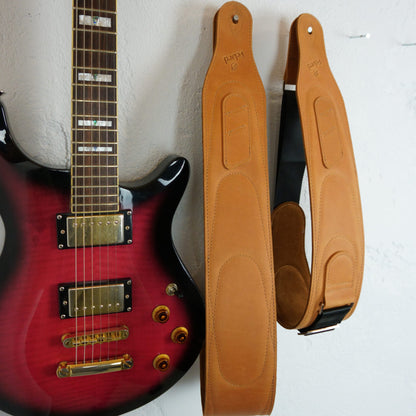A tan leather guitar straps displayed next to a red electric guitar against a white wall. The straps have stitched panels and black webbing sections with metal hardware.