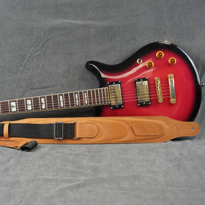 A red electric guitar with a black body finish and a tan leather guitar strap laid out on a gray textured fabric background.