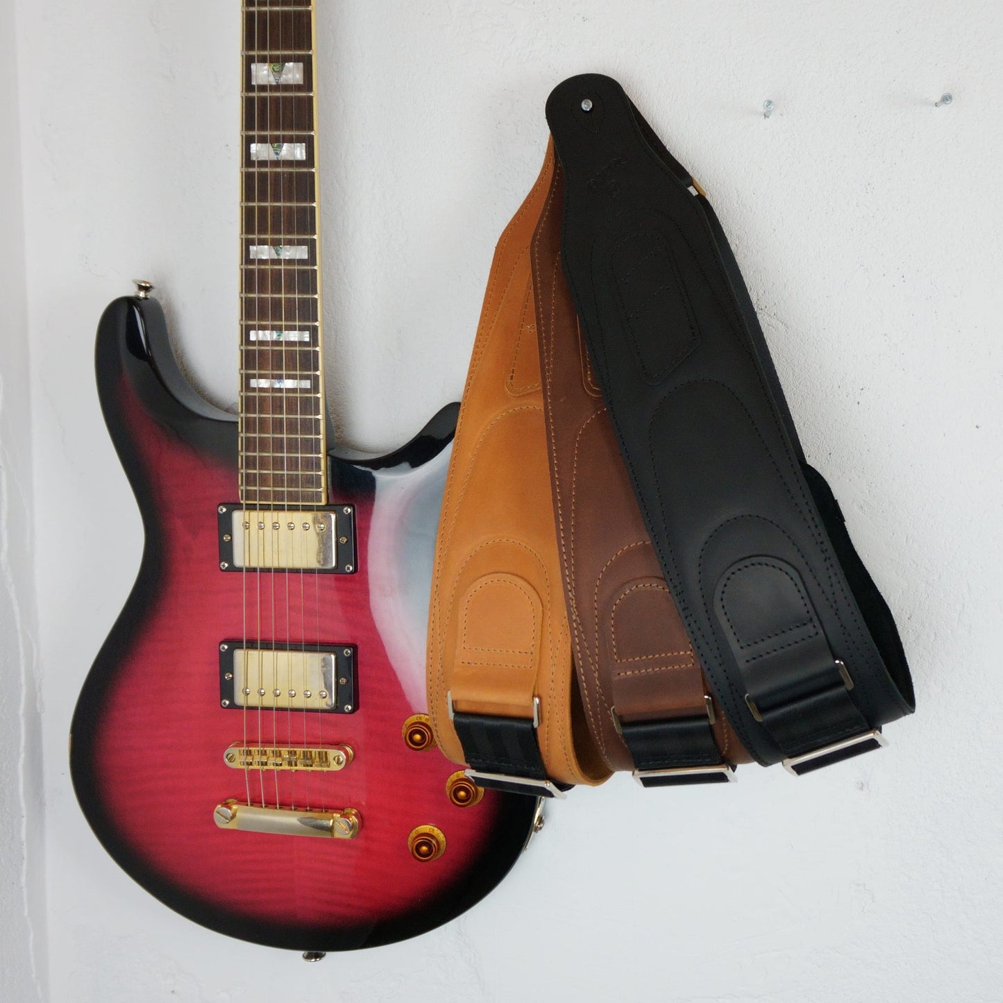 Three guitar straps in black, brown, and tan leather displayed next to a red and black electric guitar on a white wall.