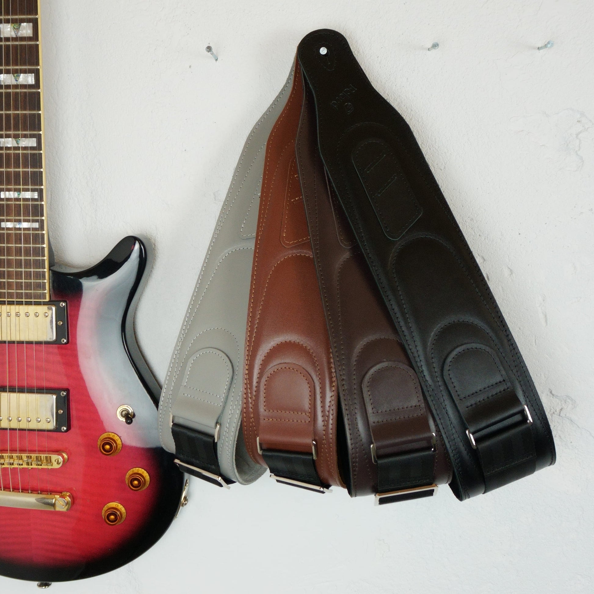 Four guitar straps in gray, brown, and black leather, arranged next to a red electric guitar on a white textured wall.
